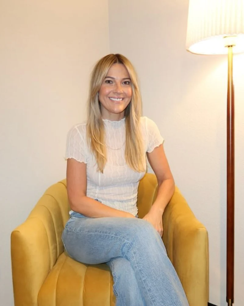 Jaime in her massage room, sitting in a mustard velvet chair beside a tall lamp